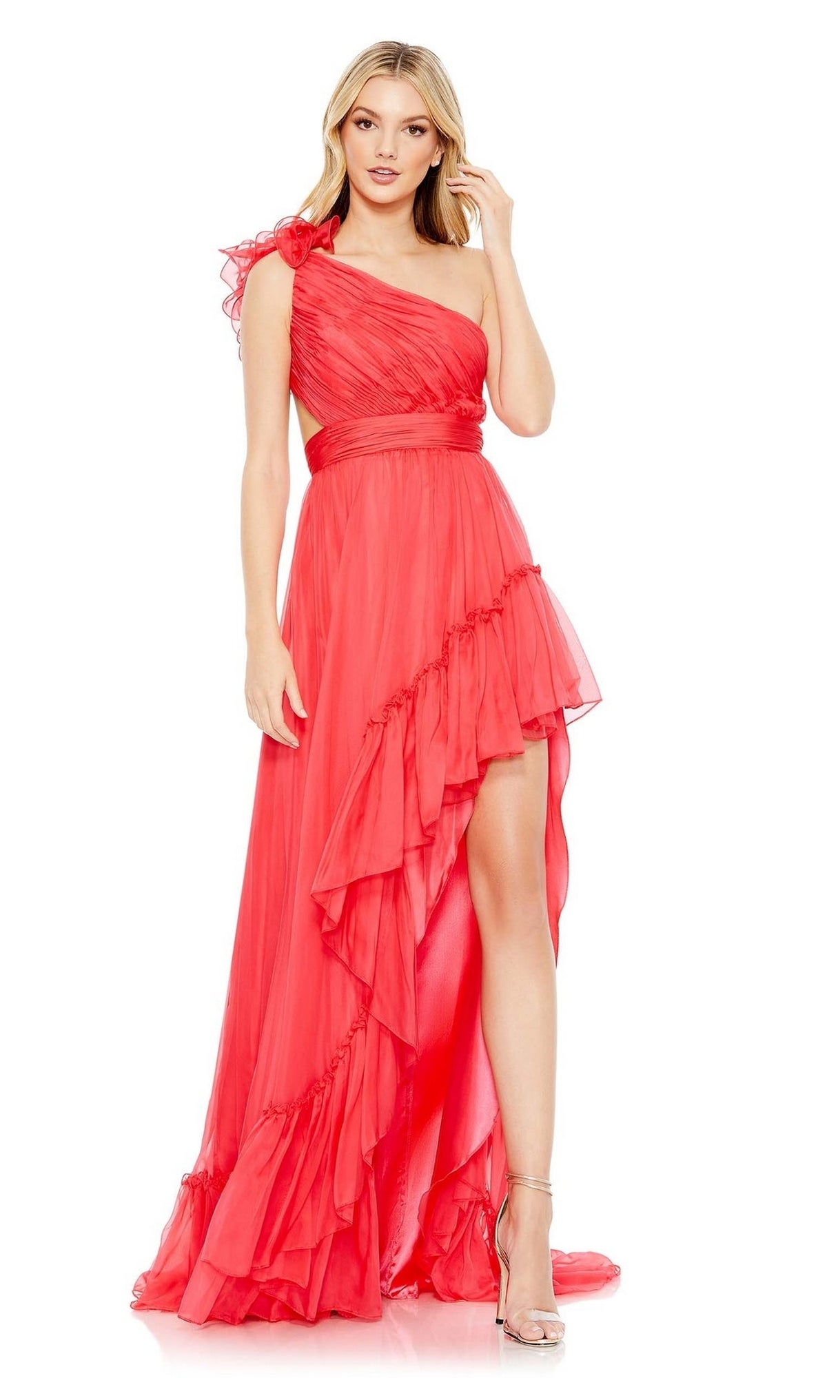 A woman wears the Long Formal Dress 67989 by Mac Duggal—a coral one-shoulder formal dress with ruffled sheer layers and a high-low hem—paired with silver heels, standing against a white background.
